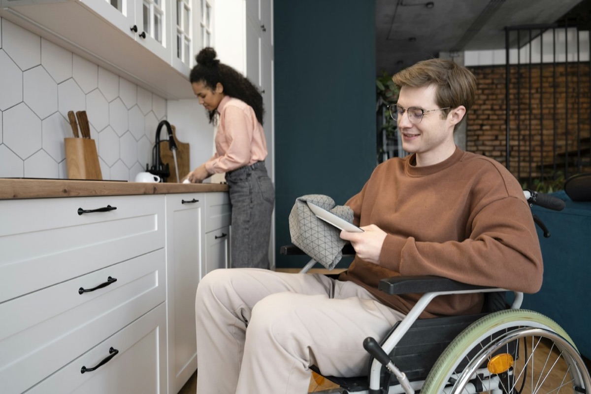 A young man in a wheelchair at home in a kitchen, with a housemate in the background